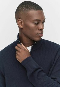 Granatowy ribbed sweter z wysokim kołnierzem i zapięciem na zamek. Posiada długie rękawy i fakturowany materiał. Widoczna biała koszulka pod spód.