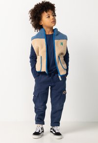 Beige und blaue Fleece-Weste mit Reißverschluss, marineblauses Langarmhemd, marineblaue Cargo-Hose und schwarz-weiße Sneakers. Mit Taschen und Logodetail.