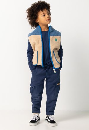 Gilet in pile zipato beige e blu, camicia a maniche lunghe blu navy, pantaloni cargo blu navy e sneakers bianche e nere. Presenta tasche e dettagli con logo.