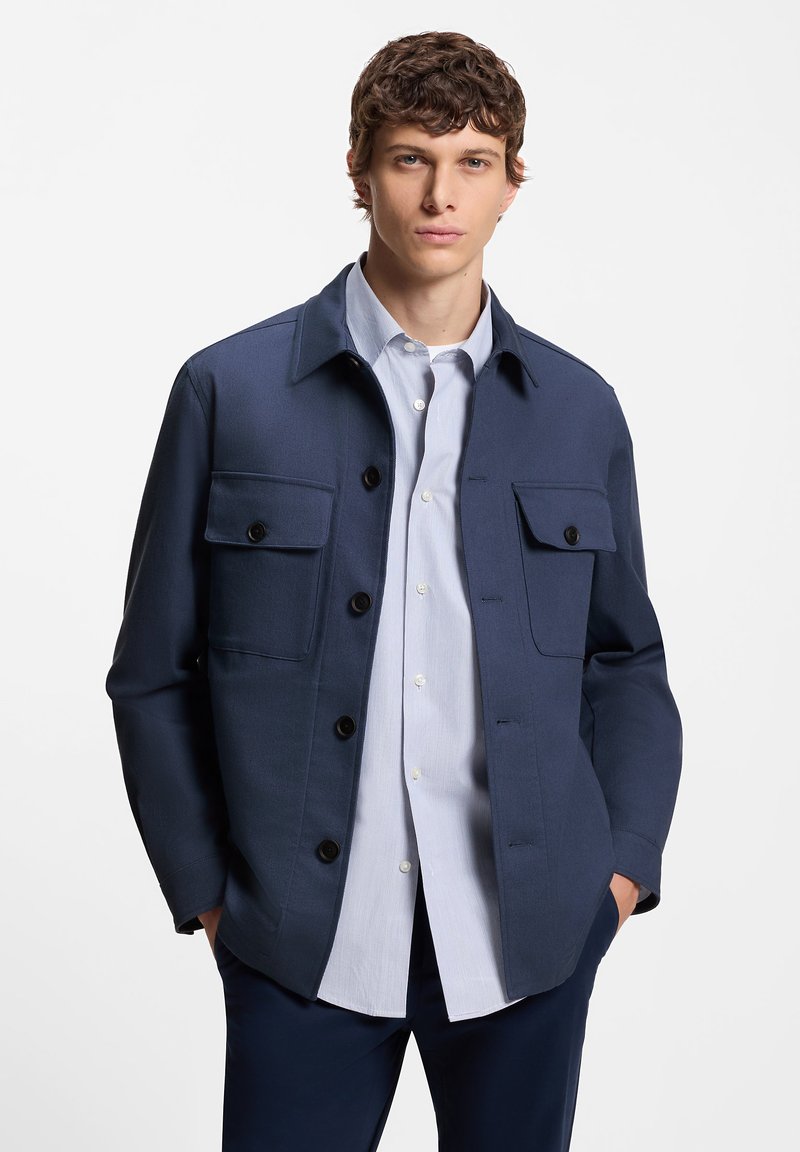 Tamsiai mėlyna striukė su keturiais priekiniais kišenėmis, užsegama sagomis priekyje ir su apykakle. Dėvima ant šviesiai mėlynos marškinių su apykakle. Lygi tekstūra.