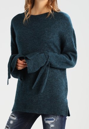 Frau trägt einen türkisfarbenen Pullover mit langen Ärmeln und verlängerten Manschetten, die zu Schleifen gebunden sind, kombiniert mit dunkelblauen Jeans im Used-Look.