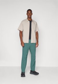 Short-sleeve beige button-up shirt over a black t-shirt, paired with green pants. Black athletic shoes complete the outfit.