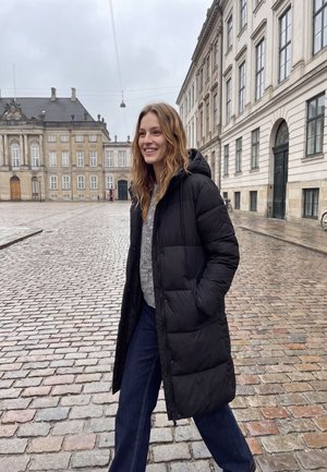Une femme souriante en long manteau noir marche à travers une place pavée bordée de bâtiments européens historiques.
