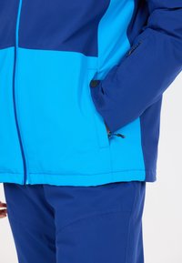 Blå och ljusblå jacka med en texturerad yta, med dragkedja och kardborre-lukning på fickorna, samt en justerbar snodd i nederkant. Byxor i matchande blå.