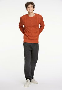 Oranges, kabelgestricktes Sweatshirt mit rundem Ausschnitt, kombiniert mit schwarzen Hosen und weißen Sneakers. Das Sweatshirt hat gerippte Bündchen und Saum.