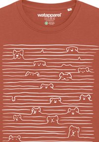 T-shirt en coton couleur rouille avec des illustrations graphiques blanches de chiens jetant un coup d'œil par-dessus des lignes ondulées, créant un motif ludique.