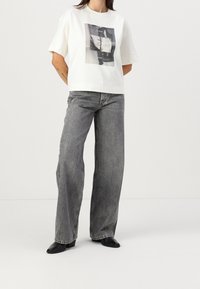 Valkoinen lyhythihainen t-paita, jossa on painettu grafiikka ja laatikkomainen istuvuus, yhdistettynä harmaisiin leveälahkeisiin farkkuihin ja mustiin korkokenkiin. Sileä pinta.