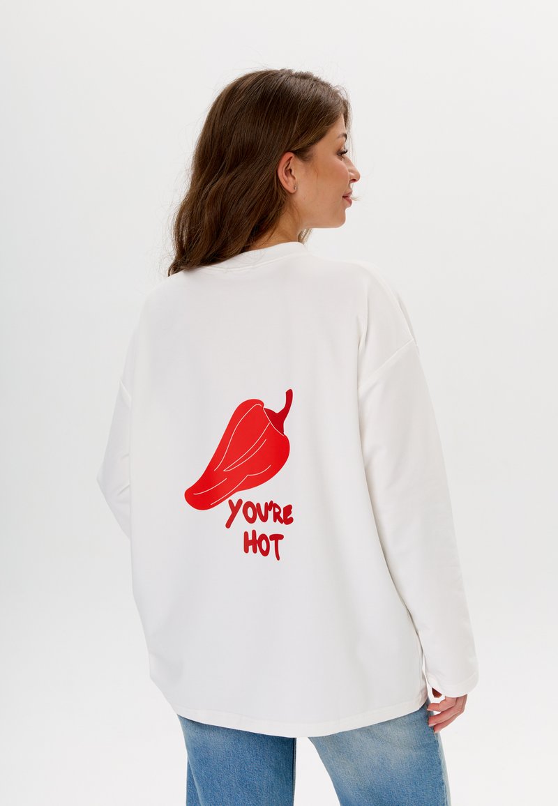 Witte oversized sweatshirt met een grafisch ontwerp van een rode chilpeper en de tekst "JE BENT HEET" in een speelse lettertype op de achterkant.