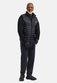 Gilet imbottito nero indossato sopra una maglietta a maniche lunghe, abbinato a pantaloni scuri e guanti. Presenta una chiusura con zip e un design elegante.