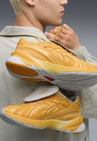 Baskets de sport jaunes avec une superposition texturée, accents blancs et semelles en caoutchouc, tenues contre un fond gris. Détail de sangle notable sur le dessus.