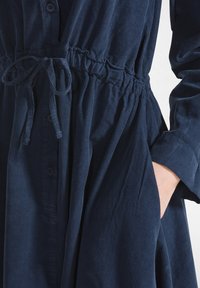 Abito in corduroy blu navy con vita stretta, chiusura a bottoni e tasche laterali. Presenta un tessuto testurizzato e maniche lunghe.