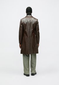 Haină maro din piele cu finisaj lucios, lungime până la genunchi și rever decupat; asortată cu pantaloni largi de culoare verde deschis și încălțăminte neagră.