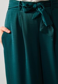 Pantalons taille haute vert canard avec un détail de nœud noué, design plissé et texture lisse ; une main dans une poche latérale.