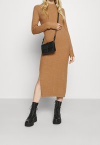 Geribbelde, lange mouw tan jurk met een zijsplit. Gedragen met zwarte combat boots en een kleine zwarte schoudertas. Eenvoudig, strak ontwerp.