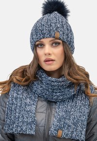 Strikket marineblå hue med en pels pom-pom og matchende tørklæde med et tekstureret mønster og et lille lædermærke til branding.