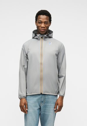 Vīrietis ģērbies pelēkā kapučjaka ar krāsainu svītrainu rāvējslēdzēju un gaiši zilām džinsu biksēm, stāvot pret baltu, vienkāršu fonu.