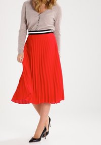 Jupe midi plissée rouge avec une ceinture à rayures noir et blanc, associée à un cardigan beige et des talons hauts noirs.