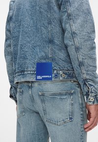 Džínová bunda a džíny ve světlém modrém odstínu, s modrou nášivkou na zadní straně s nápisem "KARL LAGERFELD JEANS" vytištěným bílou barvou.