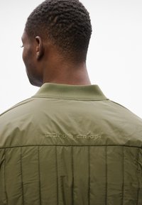 Giubbotto bomber verde oliva con texture trapuntata e colletto a coste. Presenta il logo ricamato "LINDENBERGH" sulla parte posteriore.