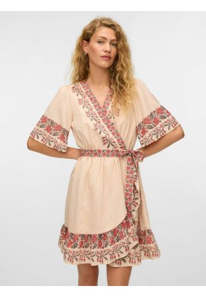 Femme aux cheveux blonds bouclés, vêtue d'une robe portefeuille beige avec une broderie florale rouge et une ceinture nouée à la taille, debout les mains sur les hanches.
