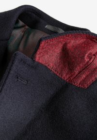 Interior de un abrigo de lana azul marino con un forro de patrón verde oscuro y un acento de bolsillo de fieltro rojo con costura detallada.