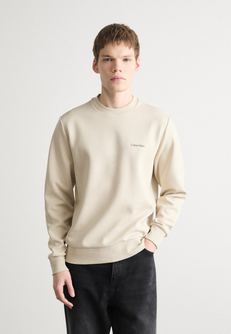 Bežová mikina s okrúhlym výstrihom a dlhými rukávmi. Na ľavej hrudi je umiestnené logo Calvin Klein. Spárované s čiernymi džínsami. Hladká textúra látky.