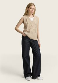 Gilet en tricot beige avec un motif en relief, design à col en V, porté sur un top blanc. Assorti à un jean large bleu marine et des baskets blanches.