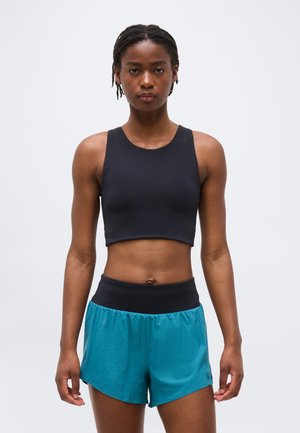 Γυναίκα όρθια, φοράει μαύρο αμάνικο crop top και πετρόλ αθλητικό σορτς με μαύρη ζώνη μέσης, έναντι απλού λευκού φόντου.