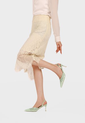 Donna con gonna midi di pizzo beige e tacchi slingback perforati verde chiaro, che solleva leggermente una gamba, su sfondo neutro.