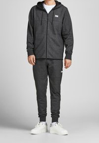 Sudadera gris con cremallera y bolsillo frontal, combinada con pantalones de chándal ajustados a juego. Zapatillas blancas con detalles en negro completan el atuendo.