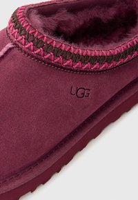 Chausson bordeaux en suedine avec une douce doublure en shearling, accents tressés rose et marron autour du col, et logo en relief.