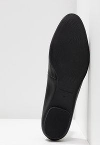 Semelle noire d'une chaussure taille 37 avec des motifs texturés antidérapants et un petit talon, positionnée verticalement contre un coin blanc.
