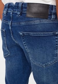 Blue denim jeans featuring a dark leather patch with "GABBA" embossed. Distressed detailing and two back pockets visible.