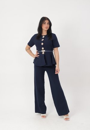 Femme portant un haut bleu marine ceinturé avec des boutons floraux et un pantalon large assorti, debout devant un fond blanc uni.