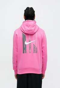 Roza kapuca dukserica s velikim bijelim Nike Swoosh logotipom i grafičkim uzorcima na leđima, izrađena od mekanog pamučnog materijala.