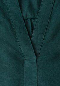 Tissu vert foncé avec un décolleté en V avec des coutures visibles et un détail froncé subtil à la couture centrale.