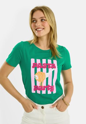 Jeune femme souriante, portant un t-shirt vert avec un motif rayé rose et le texte "Pasta party" illustrant une fourchette enroulant des spaghettis.
