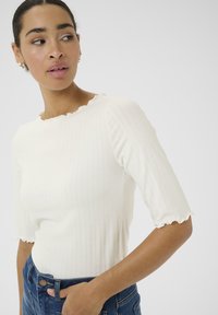 Hvid ribbede kortærmet top med en scalloped halsudskæring, tætsiddende design og blød tekstur, kombineret med blå højtaljede jeans.