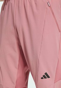 Rosa Sportshorts mit einer seitlichen Reißverschlusstasche, weicher Textur und einem schwarzen Adidas-Logo am Unterschenkel. Leichtes und atmungsaktives Material.