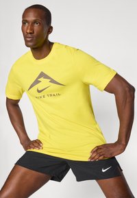 Kollane Nike Trail T-särk pruuni graafilise kolmnurga ja välgunoolise kujundusega. Kantud mustade spordilühikatega, millel on valge Nike logo.