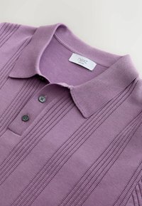 Polo violet avec un design texturé, comprenant trois boutons noirs et un col. L'étiquette indique "next" avec la date de création.