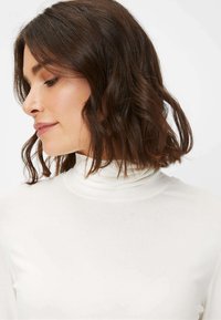 Hvid, langærmet turtleneck-top med en glat tekstur, tætsiddende design og høj krave. Håret er bølget og mellemlangt.