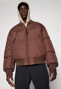 Bruine pufferjacket met een ritssluiting aan de voorkant, ribgebreide manchetten en zoom. Heeft een klein logo-opdruk. G gedragen over een lichtgekleurde hoodie.