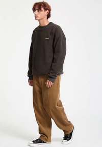 Felpa verde scuro con scollo rotondo e piccolo logo ricamato, abbinata a pantaloni di velluto a coste color tabacco e sneaker alte nere.