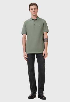 Polo shirt en coton vert clair avec un plastron à trois boutons, des manches courtes et un logo discret sur la poitrine. Associé à un jean noir et des bottes.