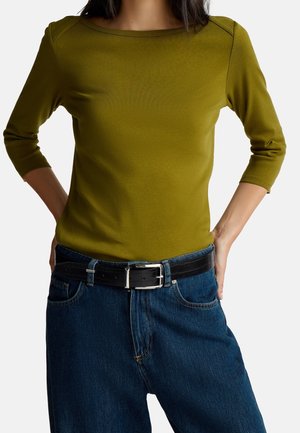 Personne portant un haut ajusté à manches longues olive avec manches 3/4 et un jean bleu taille haute avec ceinture noire.