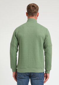 Suéter verde texturizado con cuello alto y mangas largas. Presenta puños y dobladillo acanalados, combinado con jeans de mezclilla azul.