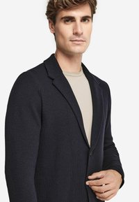 Marinblå strukturerad kavaj med en enkel knäppning, slagkrage och långa ärmar, bärs över en ljusbeige skjorta.