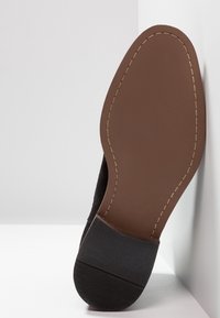 Semelle de chaussure en daim noir avec une semelle intermédiaire plate en caoutchouc marron. Présente une couture délicate le long du bord et un motif texturé pour l'adhérence.