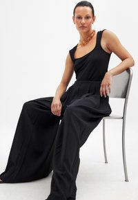 Juodas šokinėjimas su plačiomis kojomis, pristatantis prigludusią liemenį ir asimetrinį kaklą. Modelis dėvi masyvų gintaro vėrinį. Sėdi ant metalinės kėdės.